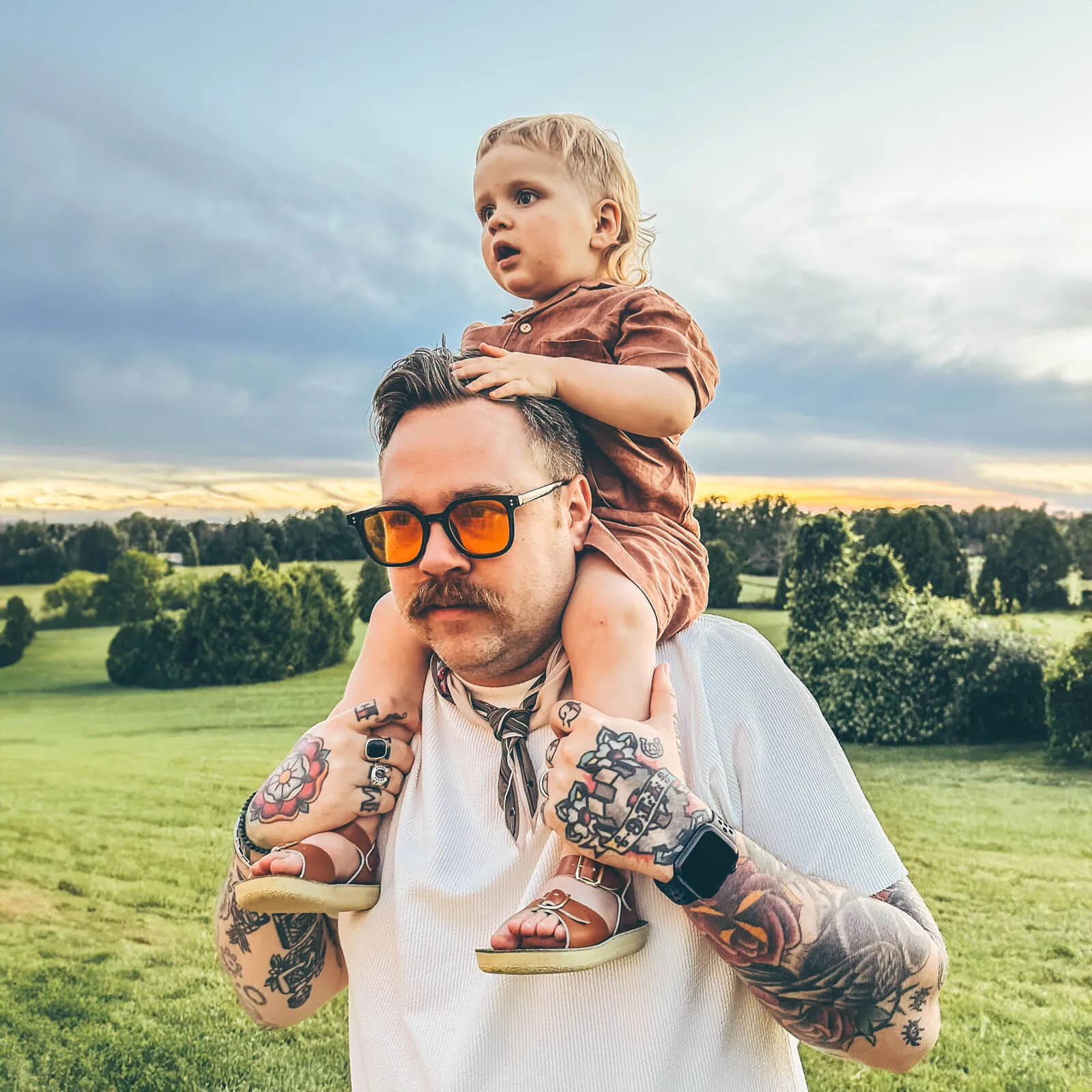 Drew Minns wearing orange-tinted glasses carrying a his son Ziggy, on his shoulders, standing in a lush, green field with a picturesque sunset in the background.