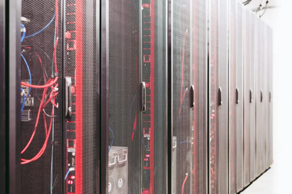 Rows of server racks in a data center, with cables and equipment visible through the mesh doors. The lighting emphasizes the organized yet complex network of red and blue cables running through the servers.