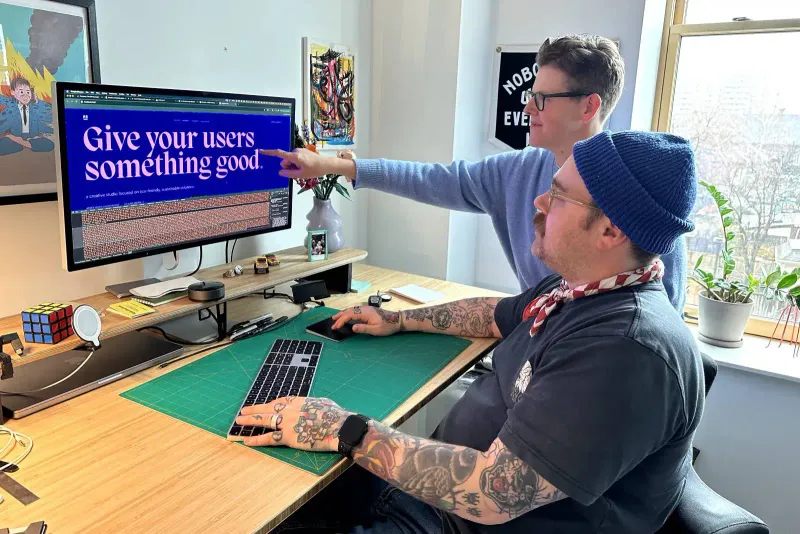 Drew Minns working with a colleague in an office, both focused on a large monitor displaying the text ‘Give your users something good.’ Drew is seated, wearing a blue beanie and a scarf, with tattoos visible on his arms. His colleague, standing beside him, points at the screen. The workspace is bright and organized, featuring a Rubik’s Cube, a vase with flowers, and various personal items. The background includes framed artwork and a window with a view of the city.