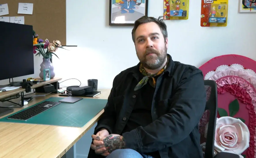 Drew Minns in his office, seated comfortably at his desk. He is wearing a black jacket and a patterned scarf, with a thoughtful expression. The desk is organized, featuring a monitor, keyboard, vase with flowers, and various personal items. The background includes framed artwork and collectible figures, creating a cozy and creative workspace atmosphere.