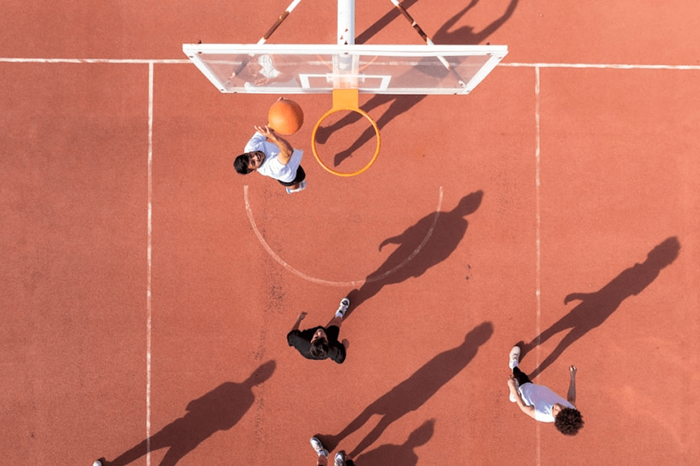 Overhead view of a basketball game on an outdoor red court, with one player mid-air about to make a layup as others look on. The scene is brightly lit with strong shadows cast by the players.