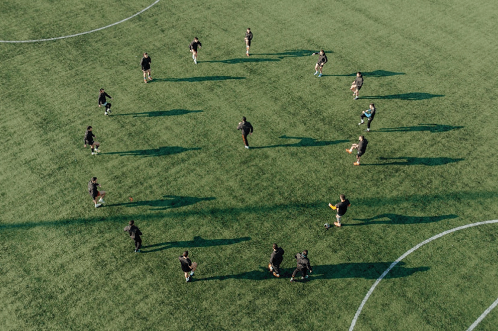 Aerial view of a group of soccer players in black training gear spread out across a green turf field, engaging in warm-up exercises or drills under bright sunlight, casting long shadows.