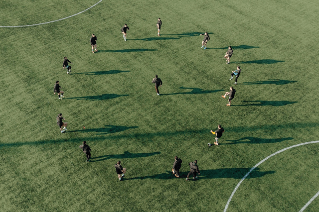 Aerial view of a group of soccer players in black training gear spread out across a green turf field, engaging in warm-up exercises or drills under bright sunlight, casting long shadows.